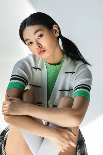 Young asian woman in striped cardigan hugging knees on grey background — Stock Photo