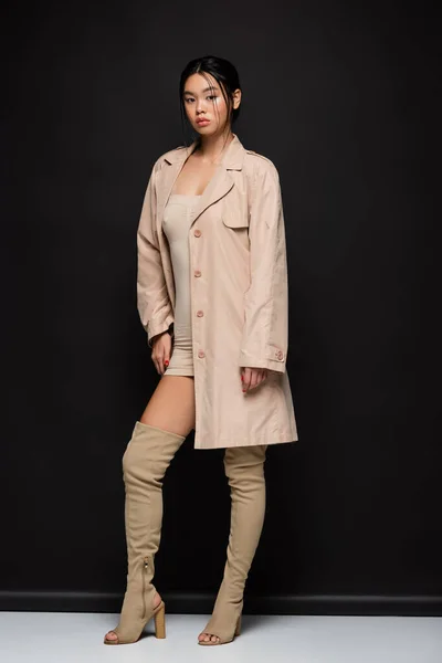 Full length of trendy asian woman in trench coat looking at camera on black background — Stock Photo