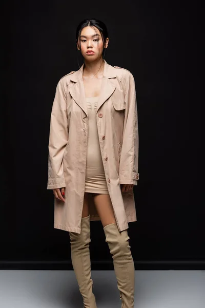 Fashionable asian model in beige trench coat looking at camera on black background — Stock Photo