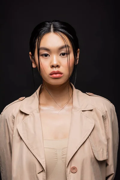 Retrato de mujer asiática en gabardina mirando a la cámara aislada en negro - foto de stock