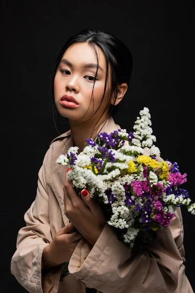 Elegante mujer asiática en gabardina sosteniendo flores silvestres aisladas en negro - foto de stock