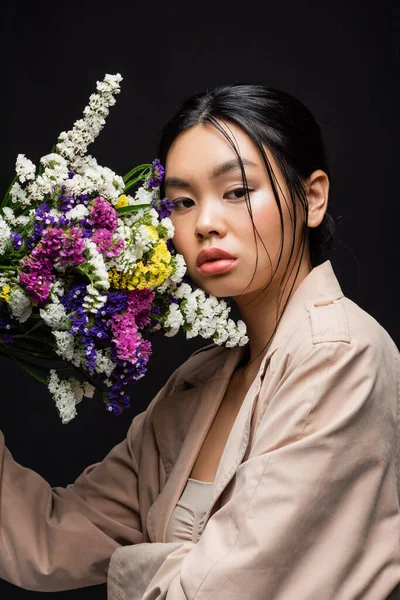 Joven modelo asiático en gabardina sosteniendo flores silvestres cerca de la cara aislado en negro - foto de stock