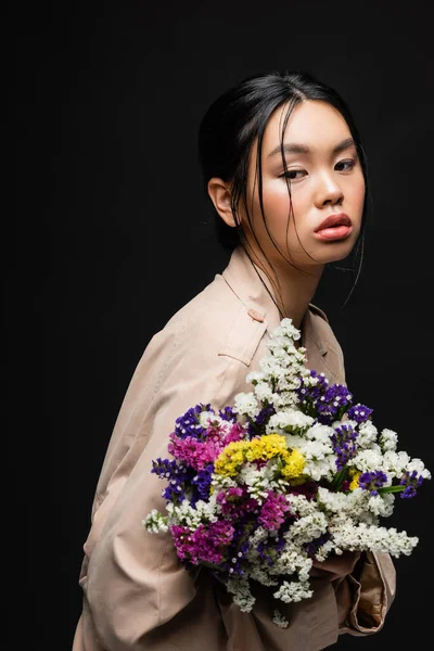 Mujer asiática en gabardina beige sosteniendo coloridas flores silvestres aisladas en negro - foto de stock