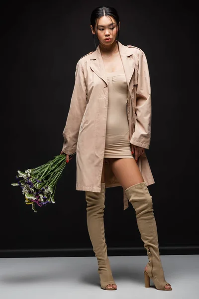 Pleine longueur de jeune femme asiatique en bottes et trench coat tenant des fleurs sauvages sur fond noir — Photo de stock