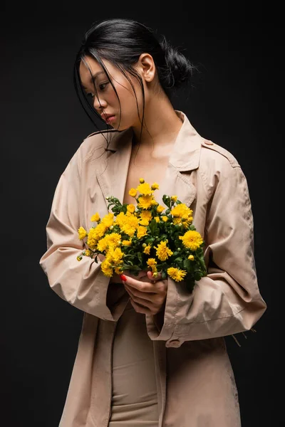 Asian model in beige trench coat holding chrysanthemums isolated on black — Stock Photo