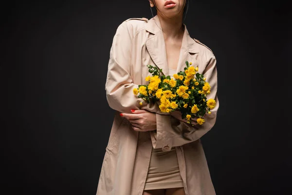 Обрізаний вид молодої жінки в траншеї пальто тримає хризантеми ізольовані на чорному — стокове фото