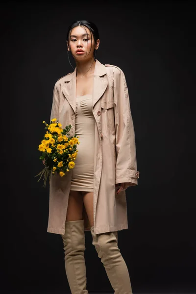 Femme asiatique en trench coat et bottes tenant des chrysanthèmes isolés sur noir — Photo de stock