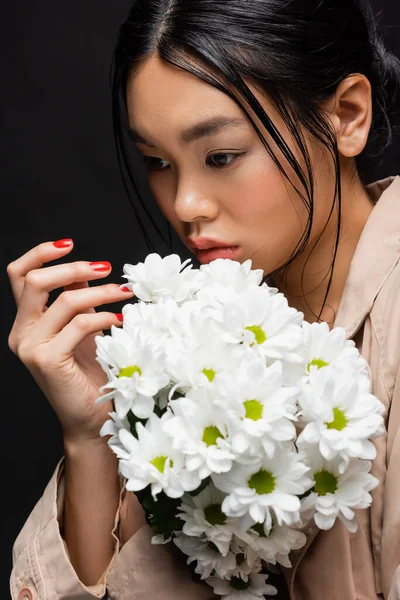 Elegante mujer asiática en gabardina posando con ramo de crisantemos aislados en negro - foto de stock