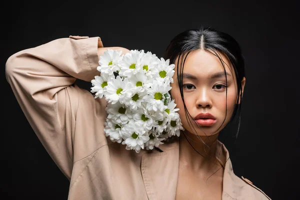 Молода азіатська модель в траншеї пальто тримає квіти біля обличчя ізольовані на чорному — стокове фото