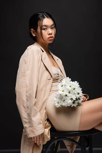 Elegante donna asiatica in abito e trench in possesso di crisantemi bianchi e seduta su sedia su sfondo nero — Foto stock