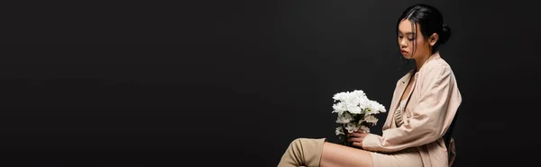 Trendy asian woman in trench coat looking at white flowers isolated on black, banner — Stock Photo