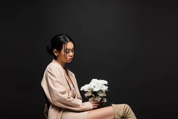 Modisches asiatisches Model in beigem Kleid und Trenchcoat mit weißen Chrysanthemen auf schwarzem Hintergrund — Stockfoto