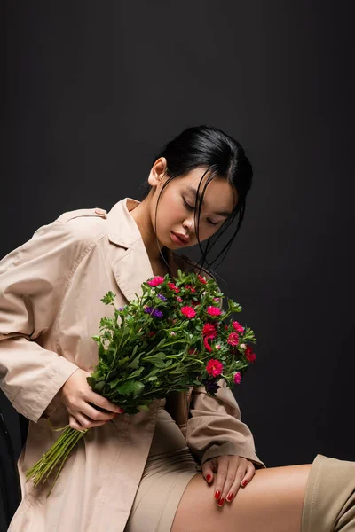 Young asian model in dress and trench coat looking at bouquet isolated on black — Stock Photo