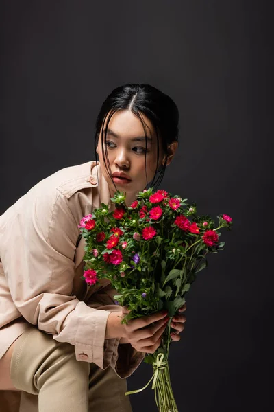 Trendy asian woman in boots and trench coat holding colorful flowers isolated on black — Stock Photo