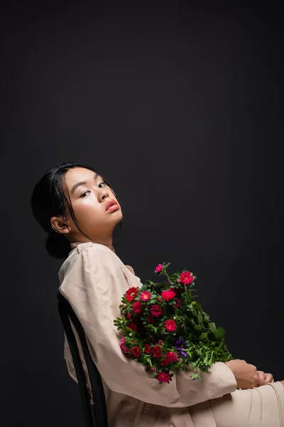 Femme asiatique élégante en trench coat et robe beige tenant des fleurs tout en étant assis sur une chaise isolée sur noir — Photo de stock