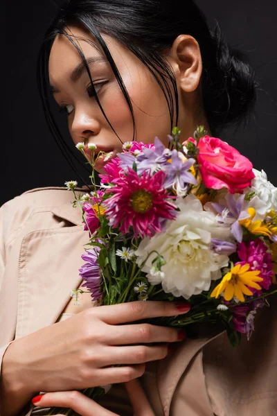 Retrato de modelo asiático en gabardina sosteniendo colorido ramo aislado en negro - foto de stock