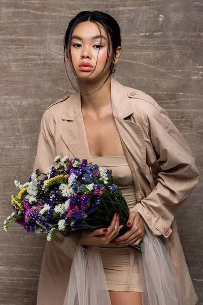 Femme asiatique à la mode en trench coat tenant des fleurs sauvages sur fond brun abstrait — Photo de stock