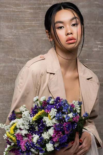 Portrait de jolie femme asiatique en trench coat tenant des fleurs sauvages sur fond brun abstrait — Photo de stock