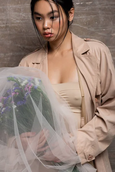 Sexy asian woman in trench coat holding bouquet under veil on abstract brown background — Stock Photo