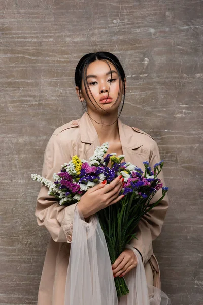 Модная азиатская модель в плаще с букетом на абстрактном коричневом фоне — стоковое фото