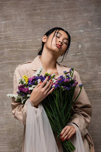 Низкий угол зрения стильной азиатской женщины в плаще держа полевые цветы на абстрактном коричневом фоне — стоковое фото