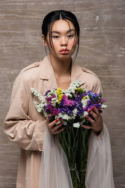 Mujer asiática de moda en gabardina sosteniendo flores silvestres de colores sobre fondo marrón abstracto - foto de stock