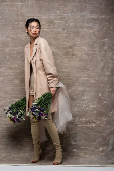 Повна довжина азіатської моделі в траншеї пальто тримає квіти з тканиною на абстрактному коричневому фоні — стокове фото