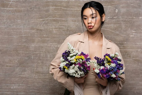 Jolie modèle asiatique en trench coat beige tenant des fleurs sauvages sur fond brun abstrait — Photo de stock