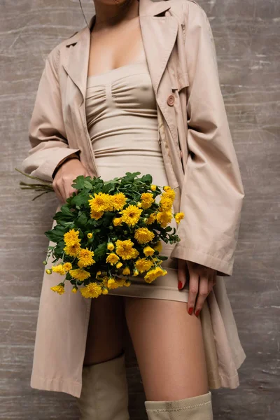 Vue recadrée d'une femme élégante en robe beige et trench coat tenant des fleurs jaunes sur fond brun abstrait — Photo de stock
