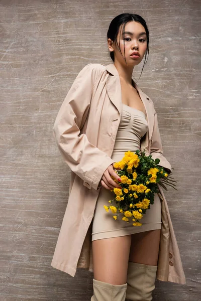 Femme asiatique élégante en robe et bottes tenant des fleurs jaunes sur fond brun abstrait — Photo de stock