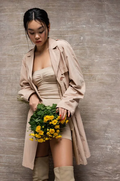 Modelo asiático elegante em casaco de trincheira tocando flores amarelas no fundo marrom abstrato — Fotografia de Stock