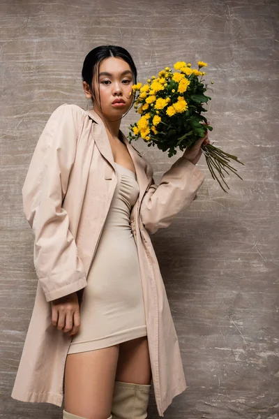 Low angle view of stylish asian woman in trench coat holding bouquet and looking at camera on abstract brown background — Stock Photo