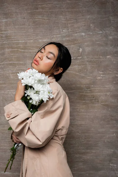 Fashionable asian woman in trench coat holding chrysanthemums and closing eyes on abstract brown background — Stock Photo