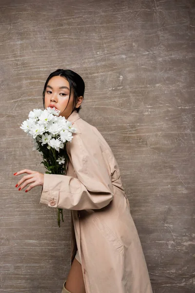 Jolie femme asiatique en trench coat posant avec des chrysanthèmes blancs sur fond brun abstrait — Photo de stock