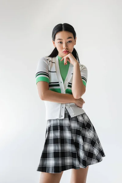 Femme asiatique élégante en jupe à carreaux regardant la caméra isolée sur gris — Photo de stock