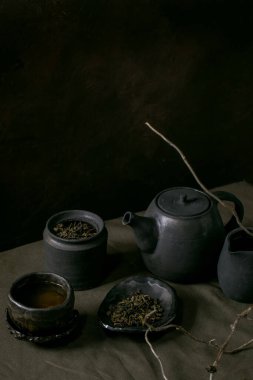 El yapımı siyah seramik çaydanlık, sürahi, kavanoz, wabi sabi bardağı, çay tabağı seti keten masa örtüsü üzerinde duruyor ve karanlık odada kuru bir dal var..
