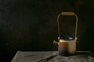 Çay seremonisi için pirinç saplı el yapımı seramik çaydanlık. Karanlık odadaki keten masa örtüsünün üzerinde duruyor..