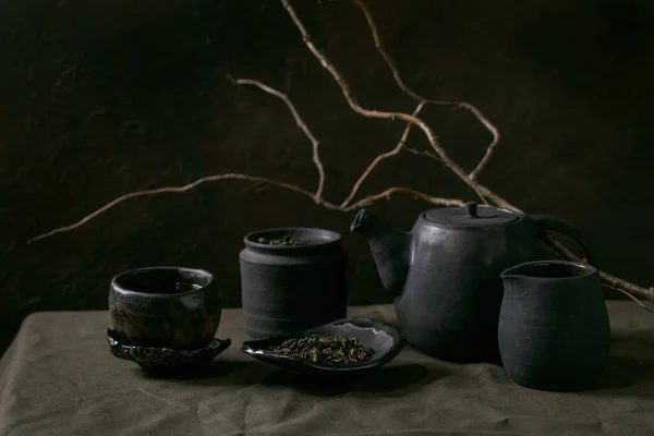 一套手工制作的黑色陶瓷茶壶壶 茶叶罐子 花生酱茶杯 茶道茶壶 放在亚麻布桌布上 深色房间有干枝条 — 图库照片
