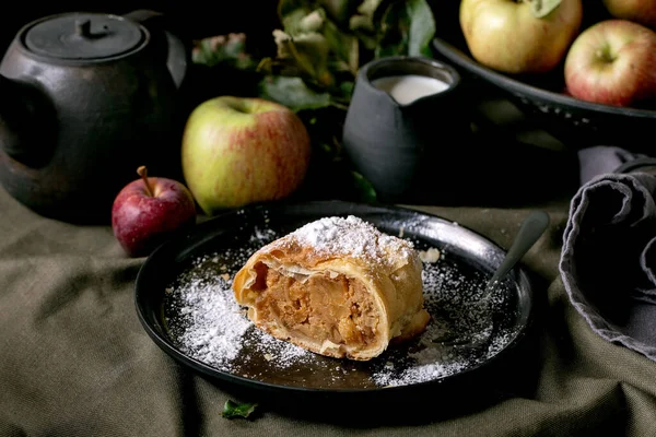 黒のセラミックプレートで手作りの伝統的なアップル ストラウデル パイのスライスは 熟した新鮮なリンゴ フォーク ダークリネンのテーブルクロスに砂糖をアイシングします ダーク素朴な秋のお茶を飲む — ストック写真
