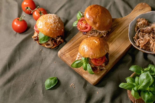 一套自制的迷你汉堡包 以炖牛肉 西红柿和罗勒配以木制切菜板和亚麻布桌布为背景 周围的杂碎和配料 现代美味快餐 — 图库照片