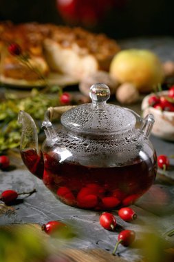 Rosehip böğürtleni, cam çaydanlıkta bitki çayı eski ahşap masa üzerinde duruyor ve yanında yabani sonbahar meyveleri ve elmalı pasta var. Sıcak vitamin içeceği.