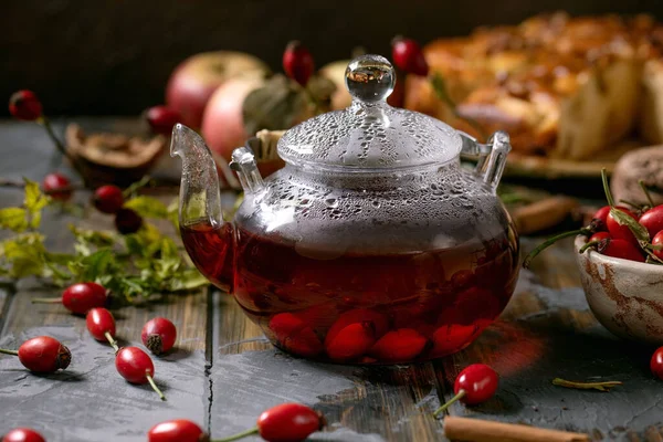 冬热饮料玫瑰草莓香草茶玻璃茶壶站在旧木板桌上 周围放着野秋浆果和苹果蛋糕 健康的热饮 — 图库照片