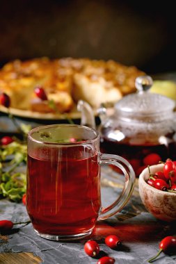 Cam bardakta gül kalçası çayı. Eski ahşap masada duruyor. Yaban güz meyveleri ve elmalı turta da var. Sağlıklı sıcak içecek..