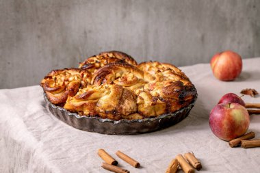 Elmalı tarçınlı çörekler yuvarlak pişirme tepsisinde duruyor. Bahçeli elmalar ve tarçın çubukları gri keten masa örtüsünde duruyor. Geleneksel ev pastanesi