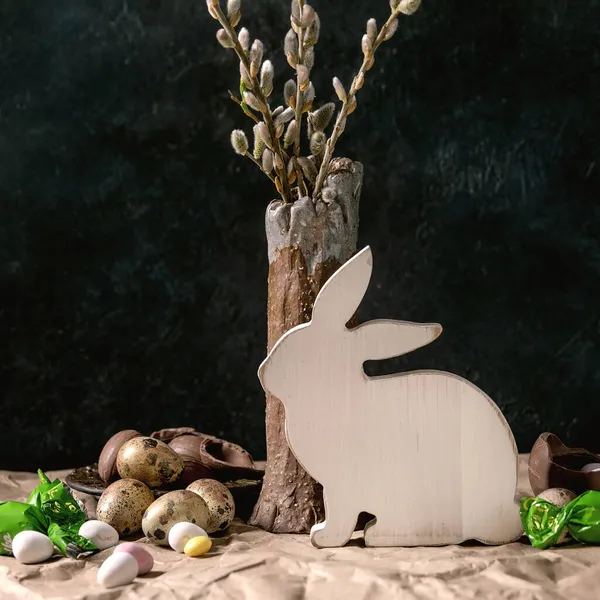陶器の花瓶 白い木製のウサギの装飾 伝統的なチョコレートの卵と紙の砕いたテーブルの上のお菓子で花柳の枝で復活祭の気分はまだ生きています 暗い背景 正方形の画像 — ストック写真
