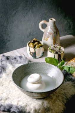 Traditional italian burrata knotted cheese in brine in grey ceramic bowl on table. Bread, olives, green salad around. Ingredients for healthy mediterranean dinner.