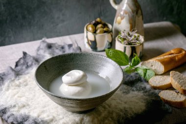Traditional italian burrata knotted cheese in grey ceramic bowl on table. Bread, olives, green salad around. Ingredients for healthy mediterranean dinner.