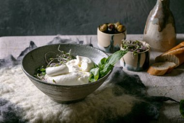 Traditional italian burrata knotted cheese salad in grey ceramic bowl on table with felted napkin. Sliced bread, olives, green sprouts around. Healthy mediterranean dinner.