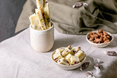 Ev yapımı beyaz çikolata, fıstıklı çikolata seramik kasede kızılcık, kareler ve şeritler beyaz keten masa örtüsünde tatlı badem. Her bayram için el yapımı yemek tatlı hediyeler..