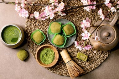 Asya pirinçli tatlı yeşil kibrit mochi, bir fincan köpüklü kibrit çayı ve yeşil kibrit tozu kahverengi masada seramik tabakta. Çaydanlık ve bahar pembe çiçekli dal örülmüş saman peçetede.
